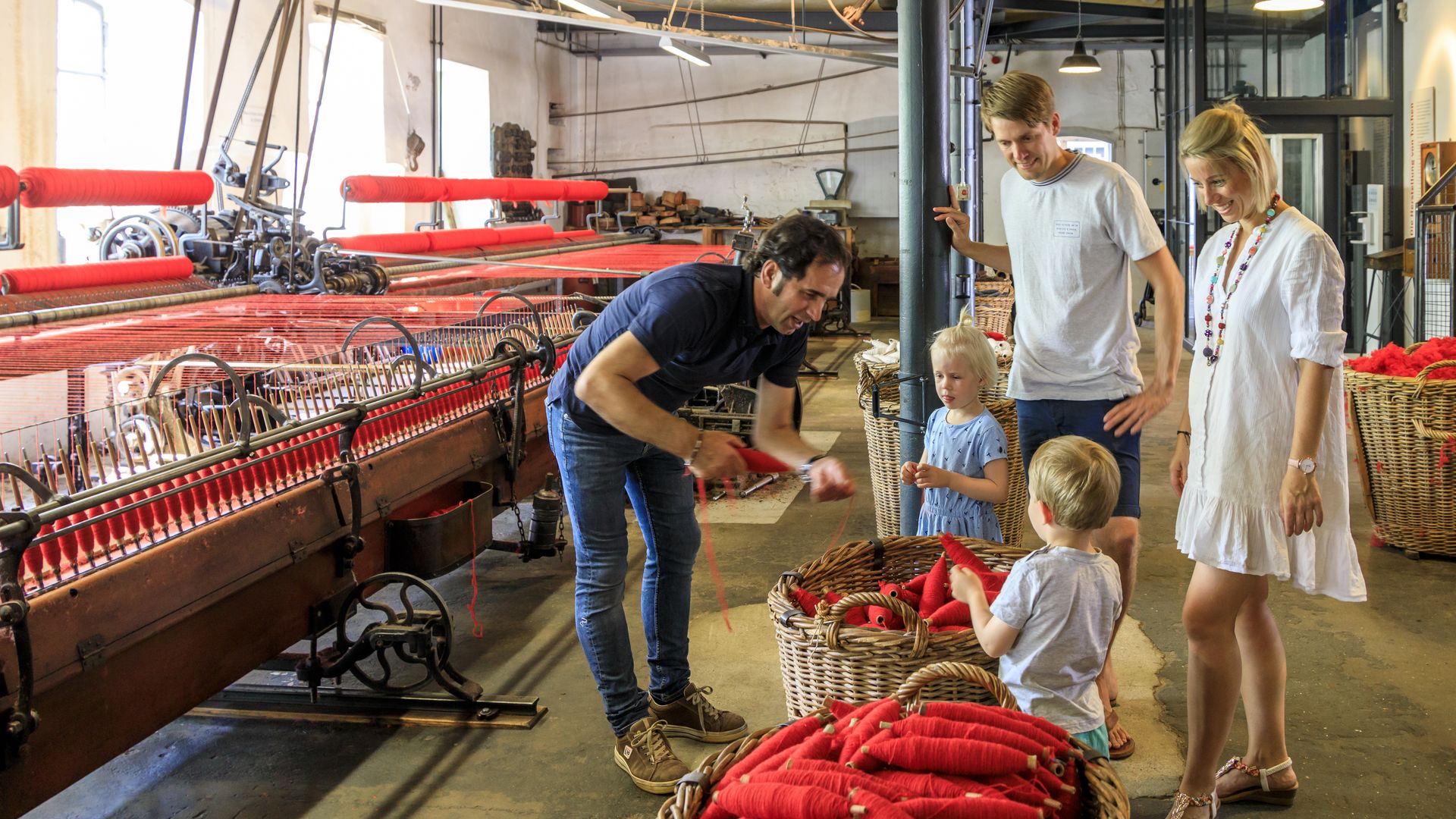 Familie besucht eine historische Weberei; Kinder schauen einem Arbeiter beim Garnspinnen zu.