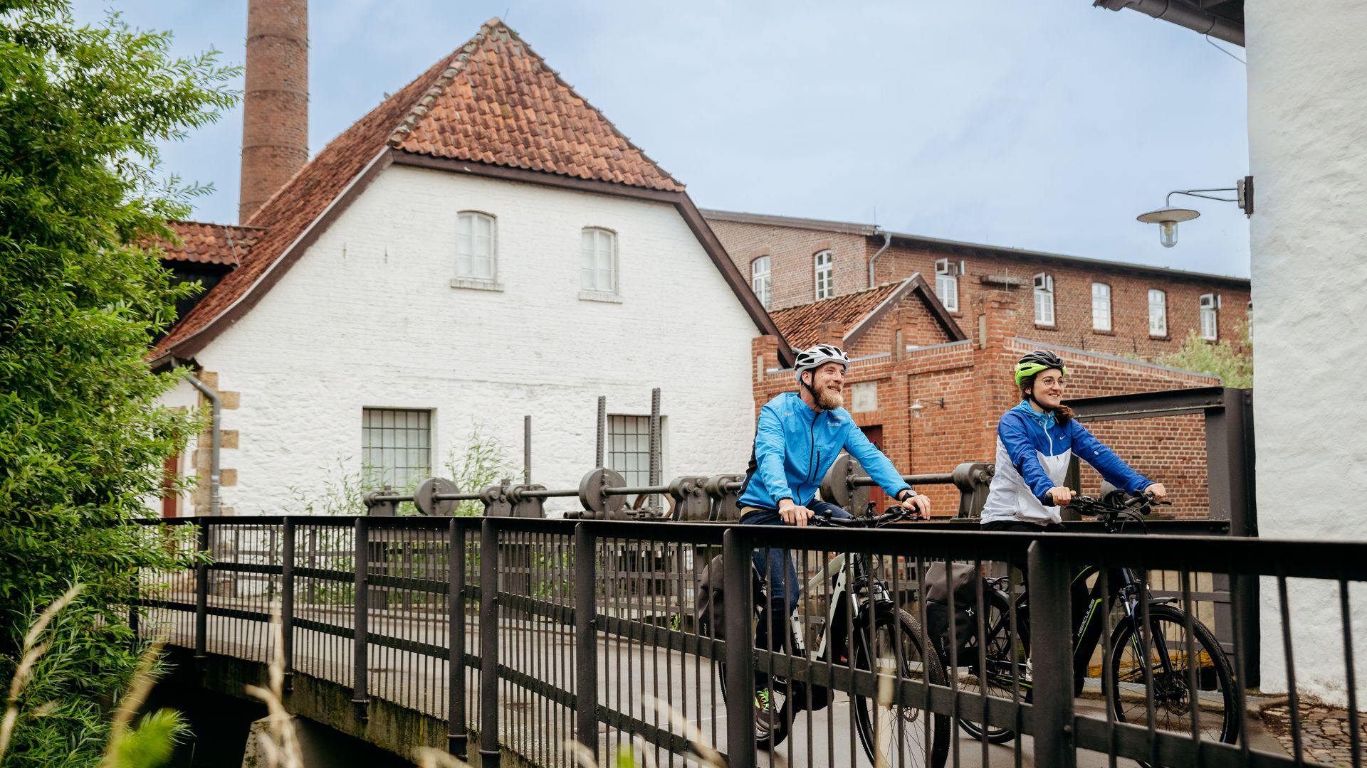 Twee fietsers steken een brug over voor een historisch gebouw met een schoorsteen.