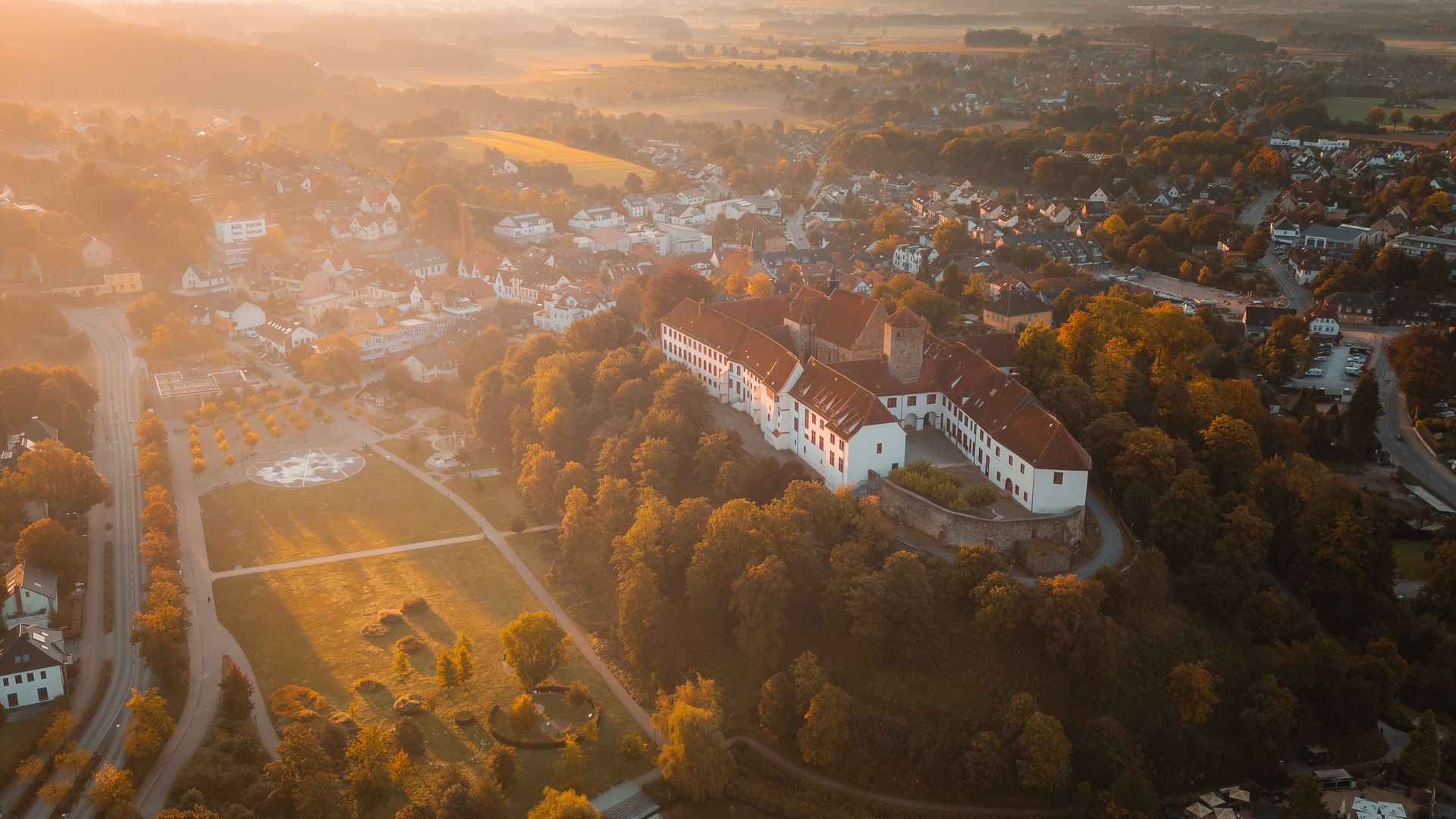 Luftaufnahme von einem historischen Schloss mit roten Dächern, umgeben von Bäumen und Wasser.