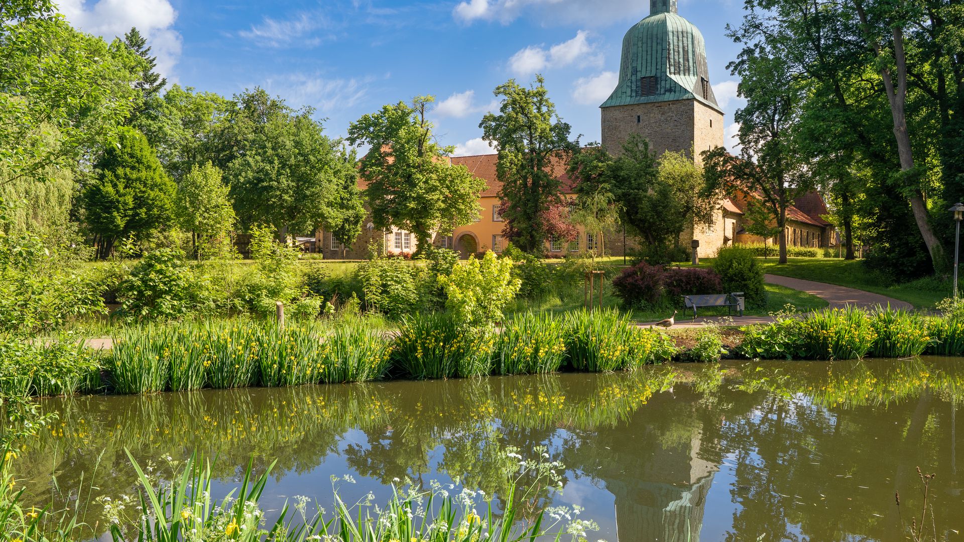 Kirchturm und Klostergebäude hinter einem Teich mit blühenden Uferpflanzen und blauen Himmel.