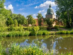 Kirchturm und Klostergebäude hinter einem Teich mit blühenden Uferpflanzen und blauen Himmel.