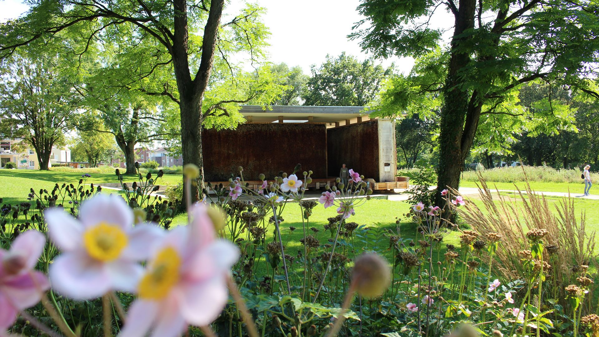 Roestkleurige stalen muur in een zonnig park, omringd door bloeiende bloemen en bomen.