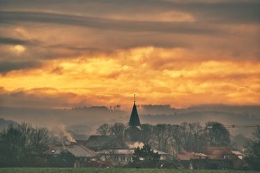 Dorf mit Kirchturm vor spektakulärem Sonnenuntergang und Nebel in der ländlichen Silhouette.