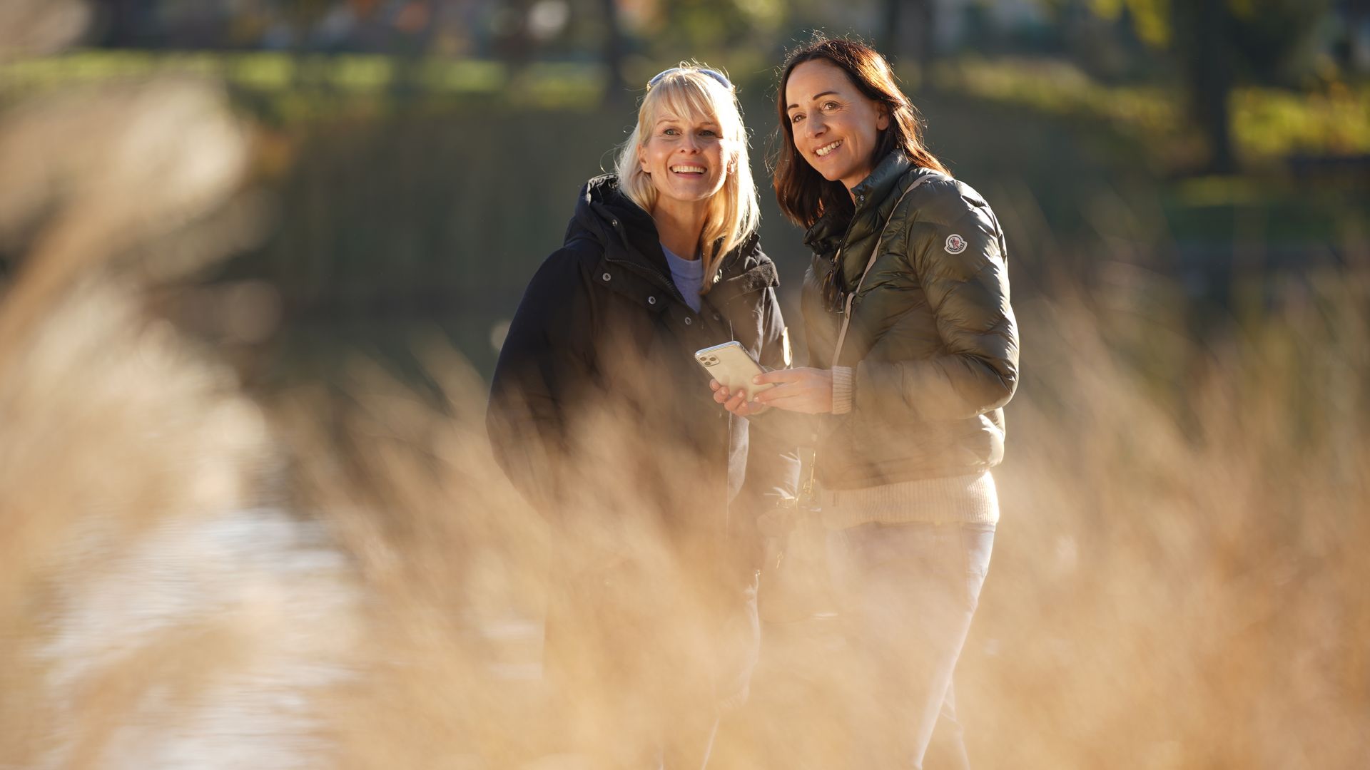 Twee vrouwen staan glimlachend in een zonnig park tegen een achtergrond van wazig gras.