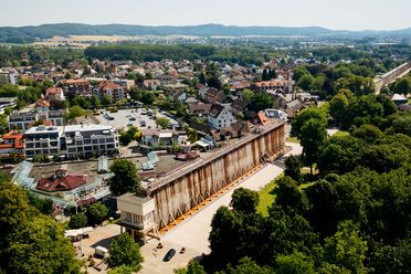 Gradierwerk in einer grünen Parklandschaft, umgeben von Stadthäusern und Hügeln im Hintergrund.