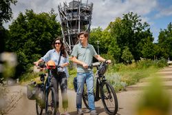 Een stel fietst glimlachend op e-bikes langs een hoge uitkijktoren.