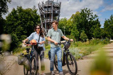 Een stel fietst glimlachend op e-bikes langs een hoge uitkijktoren.