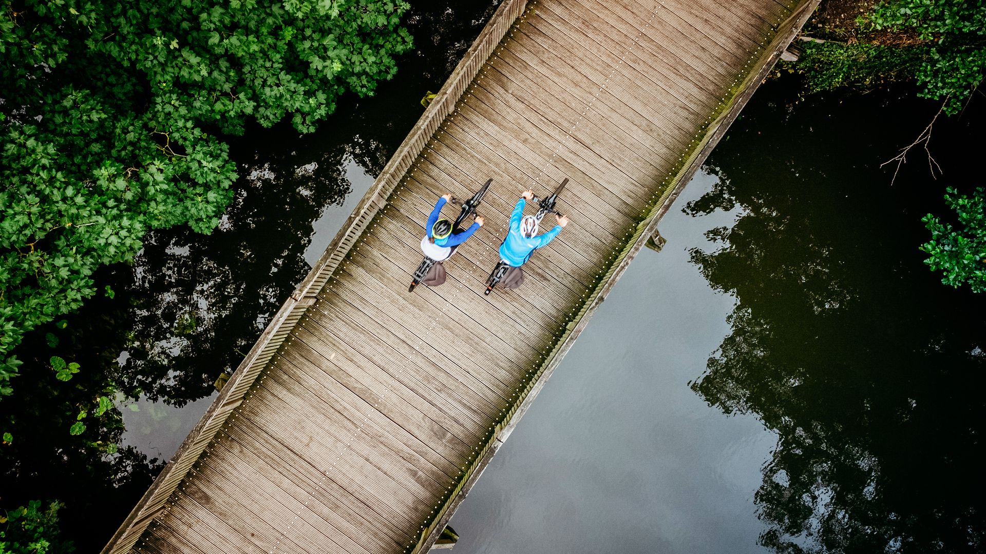 Twee fietsers op een houten brug over een rustige rivier, omringd door groene bomen.