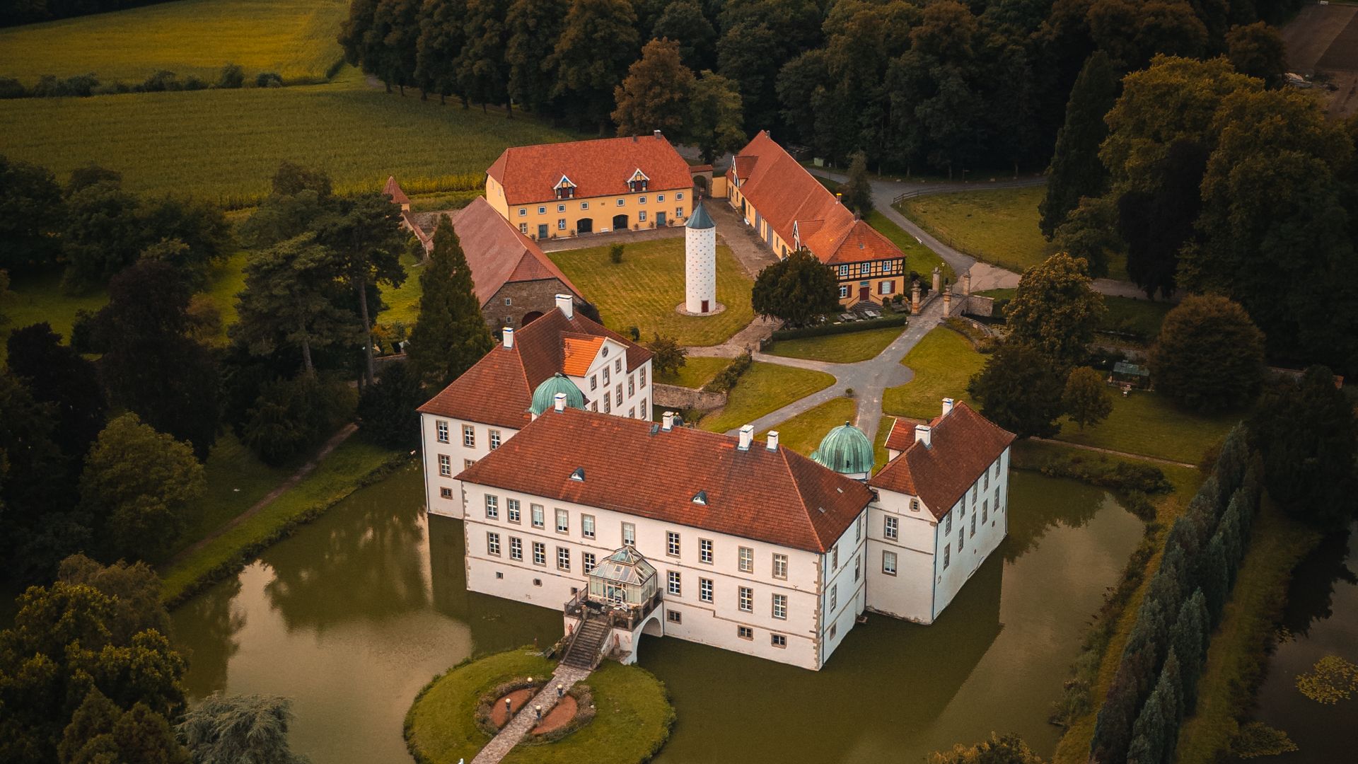Luftaufnahme eines Schlosses im Grünen mit umliegendem Wassergraben und bewölktem Himmel.