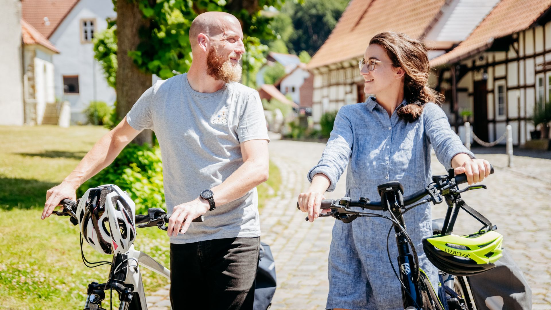 Twee mensen duwen fietsen door een zonnig dorp met vakwerkhuizen.