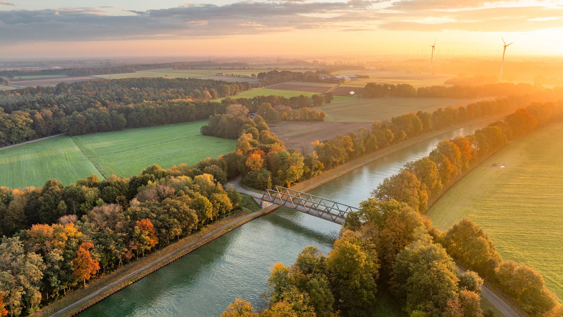 Luchtfoto van een kanaal met een brug, omringd door herfstkleurige bomen bij zonsondergang.
