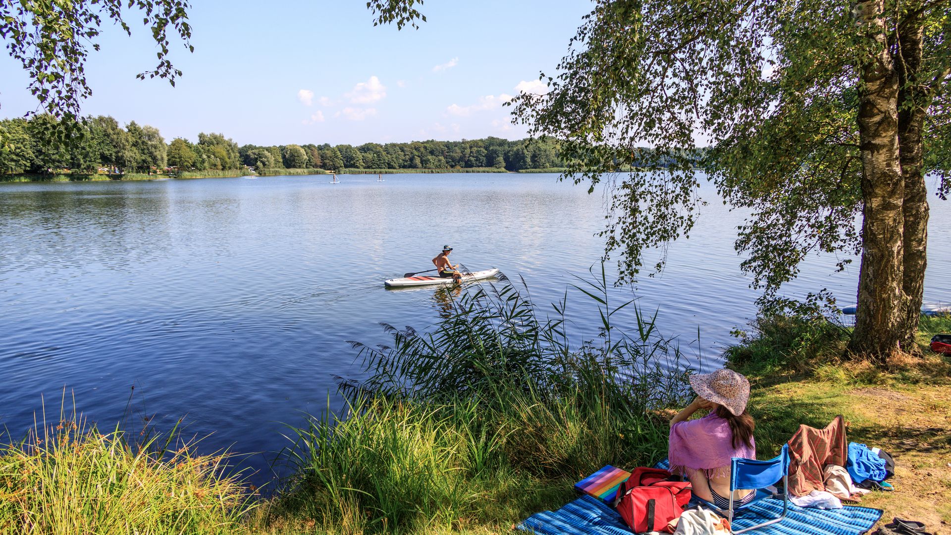 Ein ruhiger See mit einem Paddelboarder und einer Person am Ufer auf einer Picknickdecke.