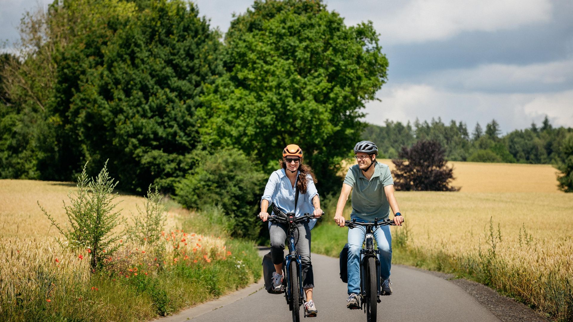 Zwei Radfahrer auf einem ländlichen Weg, umgeben von Feldern und Bäumen.