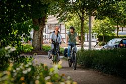 Twee fietsers rijden over een schaduwrijk pad door Bad Laer, omgeven door groene ruimten en stedelijke bebouwing.