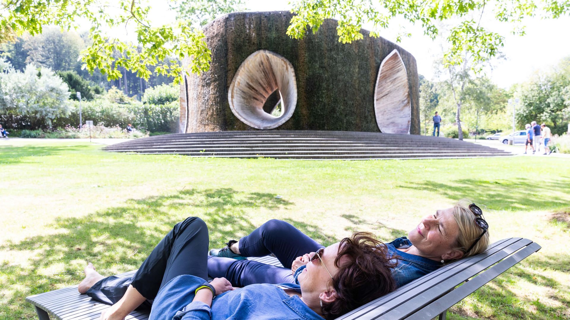 Twee vrouwen relaxen op een bankje voor een grote, sculpturale fontein in het park.