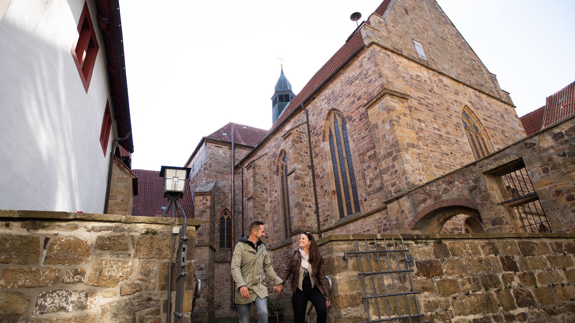 Echtpaar aan het wandelen in een oud steegje, geflankeerd door historische stenen huizen en een kerk op de achtergrond.