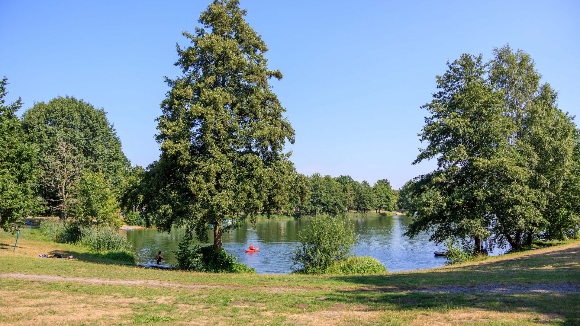 Een groen park met een meer, omringd door bomen; twee mensen peddelen op het water.