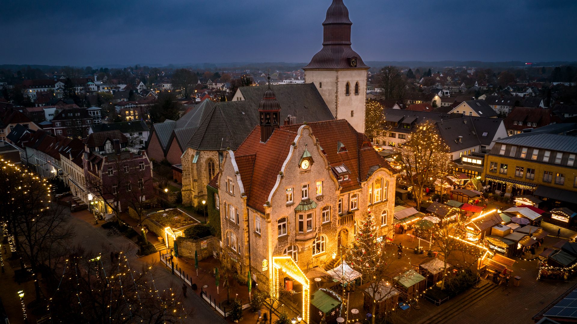Avondlijk stadsgezicht met verlichte kerstmarkt en historische kerk op de achtergrond.
