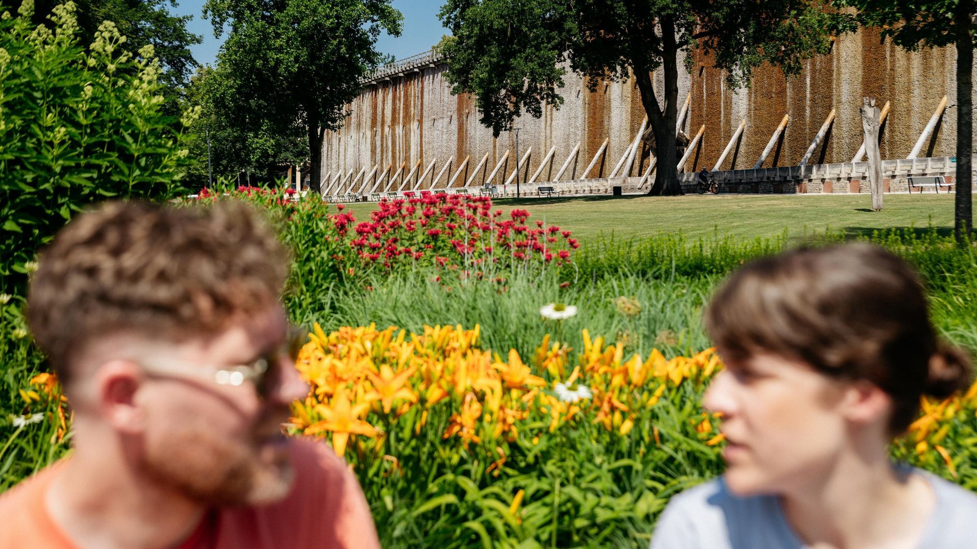 Twee personen voor een oud gradierwerk, met op de voorgrond een bloeiende tuin.